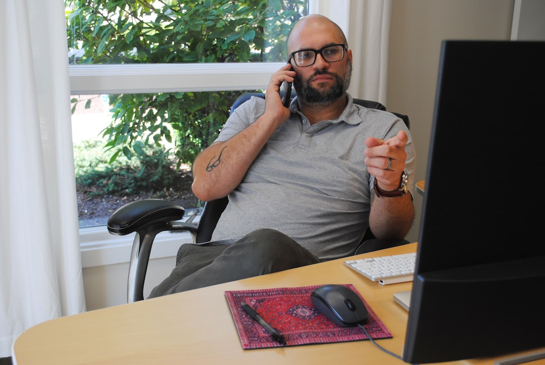 A man with glasses and a gray polo shirt, seated at a desk, is holding a phone to his ear while pointing with his other hand. A computer monitor and a wireless keyboard are visible on the desk along with a mouse and a decorative mat.