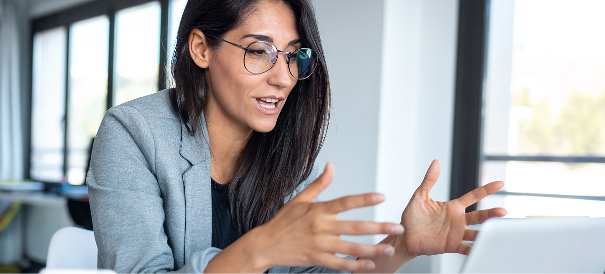 A woman in glasses and a grey blazer gestures with her hands while speaking to someone during a video call, seated indoors near large windows.
