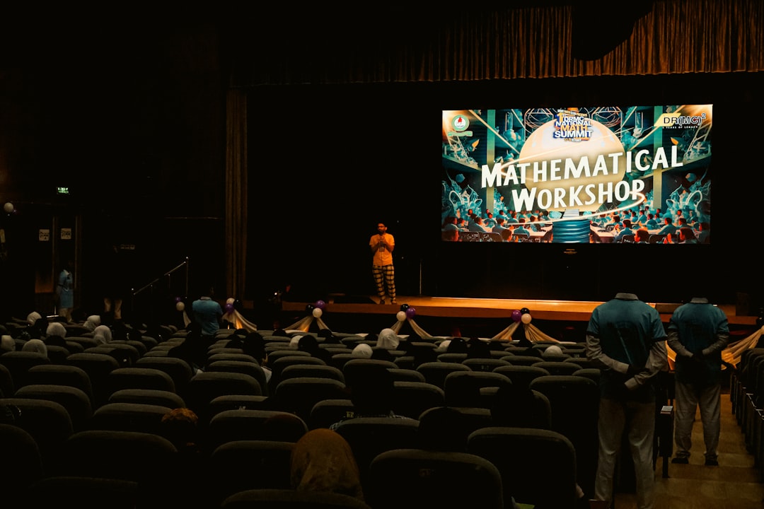 An auditorium set up for a workshop, featuring a large screen displaying "MATHEMATICAL WORKSHOP." A speaker in orange stands on stage, while attendees, mostly in headscarves, are seated in rows of chairs facing the stage.